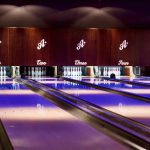 Interior view of the bowling alleys at All Star Lanes bowling alley, Westfield Stratford, East London. Lighting is pink and purple upon light wood bowling lanes with white bowling pins stood ready to play; lanes are marked with the branding "A*" and numbered one to four.