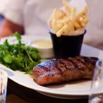 Medium rare steak, with side serving of green watercress and black pot of French fries styled in a white paper cone, as served on an oval white plate at Côte Brasserie, Barbican Centre branch in the City of London. In soft focus around the dish lies a candle in a tealight holder, a glass of rosé wine and a small dish of Béarnaise sauce. The backdrop is that of a man in a white short-sleeved shirt.